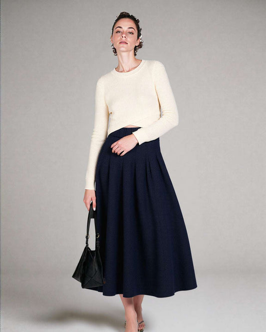 Woman wearing a white sweater and navy skirt holding a black handbag against a plain background