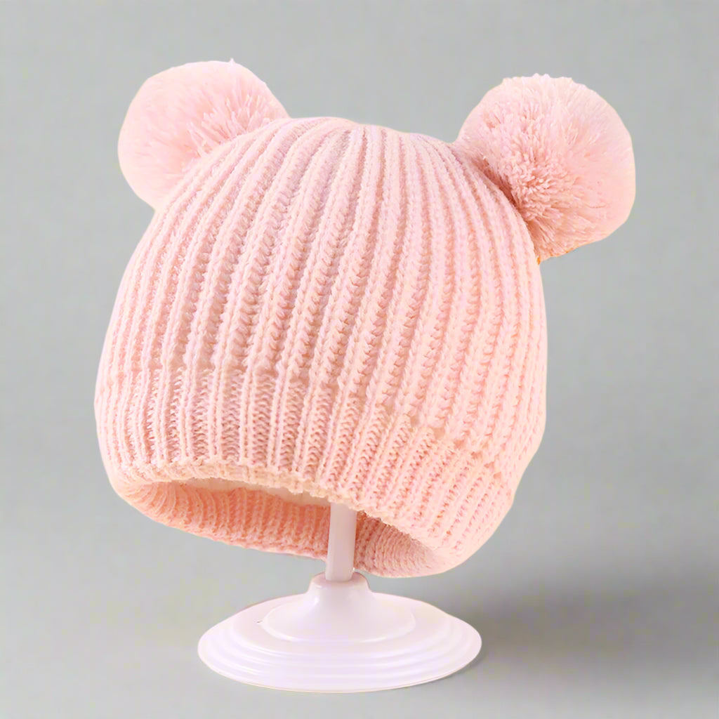Pink knitted hat with pom-poms on a stand against a light yellow background