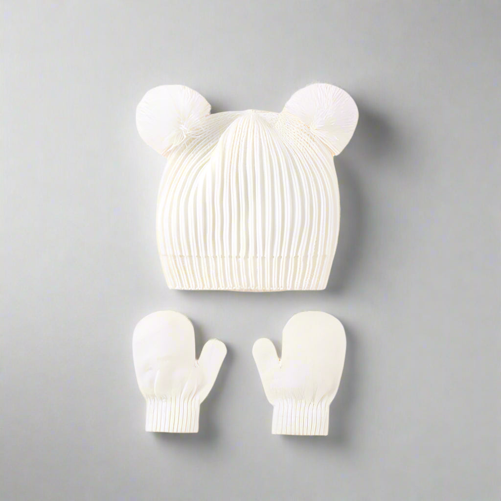 White knit baby hat with pom-poms and matching mittens on a gray background