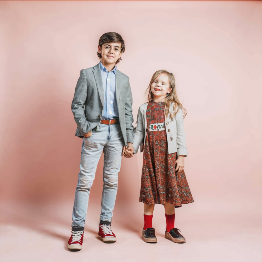 A young boy and a girl posing together while holding hands for children's clothing collection standing against a light pink studio setting background