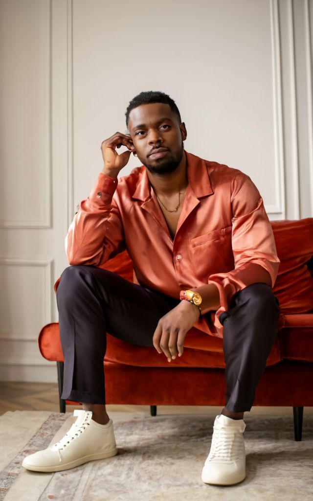 An African American man siting on a sofa dressed in romantic style satin oversized long sleeved shirt, black suit pants and white sneakers. Looking forward to a date night.