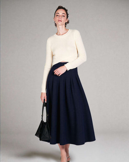 Woman wearing a white sweater and navy skirt holding a black handbag against a plain background