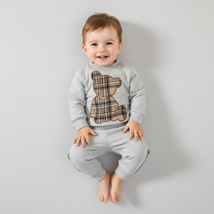 Baby wearing a gray outfit with a teddy bear design, surrounded by toys and decorative items on a light background.