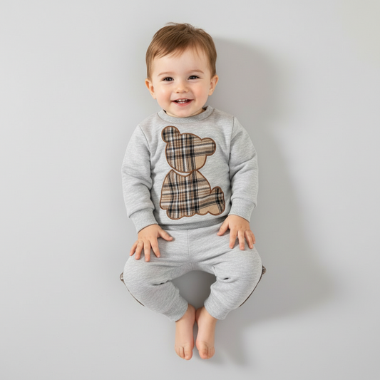 Baby wearing a gray outfit with a teddy bear design, surrounded by toys and decorative items on a light background.