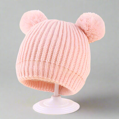 Pink knitted hat with pom-poms on a stand against a light yellow background