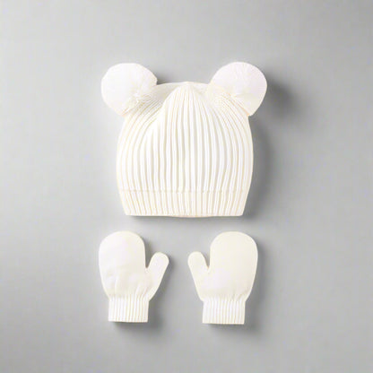 White knit baby hat with pom-poms and matching mittens on a gray background